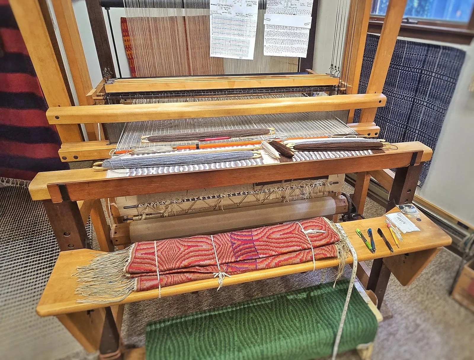The warped loom in Charles H. Blanchard's Dyberry Weaver studio in Narrowsburg NY, shuttles resting mid-weave with a finished red hand-woven piece on the bench